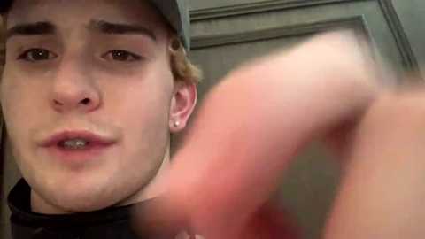 Media: Video of a young, fair-skinned male with light brown hair and a cap, holding a phone, with a blurred background of a door and wall.