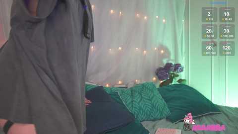 Media: Video of a woman in a gray t-shirt, standing beside a bed with green pillows, surrounded by string lights and flowers.