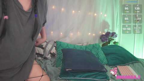 Media: Video of a person in a dark shirt and black stockings, standing next to a bed with green pillows and a camo blanket, in a softly lit room with fairy lights and a bouquet of purple flowers.