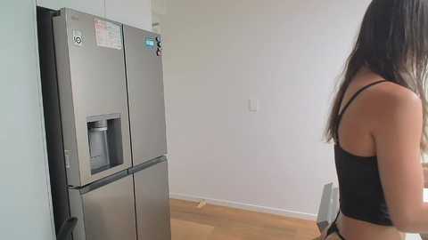 Media: Video of a young woman with long, straight, dark hair, wearing a black tank top, standing in a minimalist, modern kitchen with a stainless steel fridge and light wooden floor.