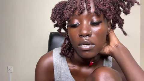 Media: Video of a Black woman with a deep brown complexion and natural, shoulder-length curly hair, wearing a grey tank top. She has a serious expression, with her head resting on her hand. Background is a plain, beige wall with a power outlet visible.