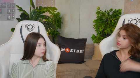 Video of two young women in gaming chairs, one with long dark hair, one with wavy brown hair, in a cozy room with green plants and a black pillow.