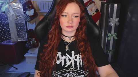 Media: Video of a fair-skinned, long-haired redheaded girl in a black \"Ondskapt\" t-shirt, seated in a black gaming chair, surrounded by holiday decorations and gift-wrapped presents in a dimly lit room.