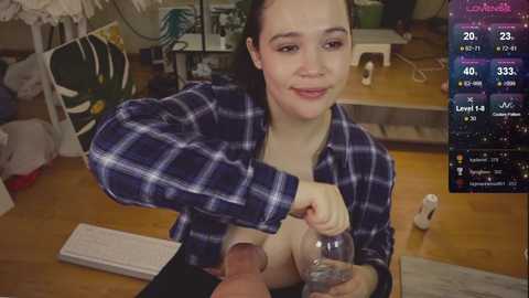 Media: A video of a young woman with pale skin and dark hair, wearing a blue plaid shirt, sitting at a wooden table, holding a glass jar. Background includes a TV with streaming stats, a laptop, and clutter.