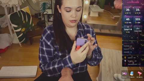 Video of a young woman with long black hair, wearing a blue plaid shirt, using a smartphone, seated on a wooden floor in a cluttered room with a green bag and a chair in the background.