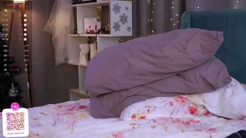 Media: Video of a cozy bedroom with a purple pillow and white floral duvet on a bed, soft lighting, white shelves with decor, and a teal headboard in the background.