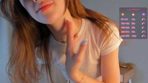 Media: Video of a young woman with long, light brown hair, fair skin, and a slender physique. She wears a white T-shirt, touching her neck, with a social media profile overlay in the background.