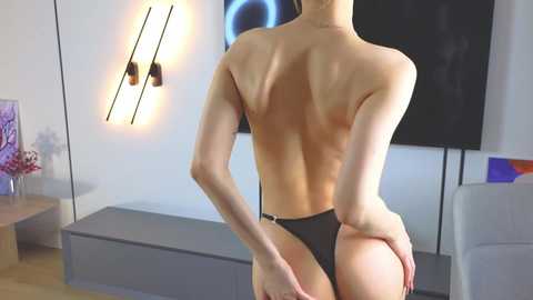 Media: Video of a slender, fair-skinned woman with a toned back, wearing black thong panties, standing in a modern living room with minimalistic decor, including a black TV and a colorful abstract painting.