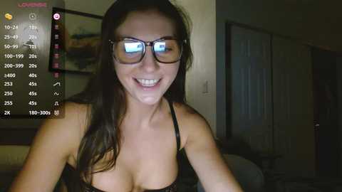 Media: Video of a smiling woman with long brown hair, wearing glasses and a black lace bra, captured in a dimly-lit room with a digital clock on the left.