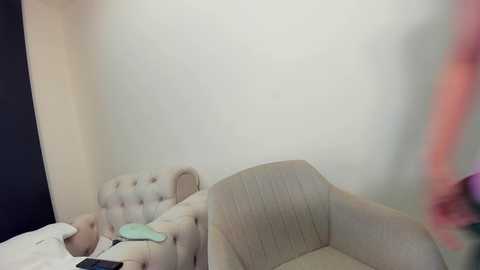 Media: Video of a beige, tufted armchair in a minimalist room with a plain white wall and a blurred figure in pink clothing.