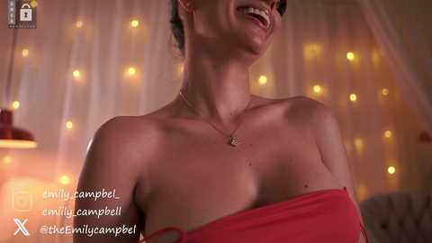 Video of a smiling, light-skinned woman with dark hair, wearing a red off-shoulder top, in a softly lit, cozy room with fairy lights and draped fabric.