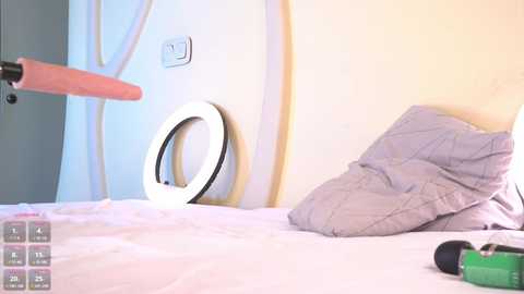 Media: Video of a bed with a white sheet, a gray pillow, a green camera, and a ring light mounted on the wall, featuring a large, erect, pink dildo.