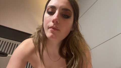 Media: Video of a young woman with light skin, long brown hair, and dark eyeliner, wearing a strapless top, leaning forward slightly, indoors with white walls and a radiator.