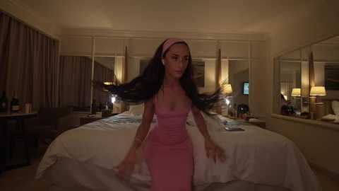 Media: Video of a woman with long dark hair, wearing a pink dress, dancing in a dimly lit hotel room with a large bed, mirrored wardrobe, and lamps.