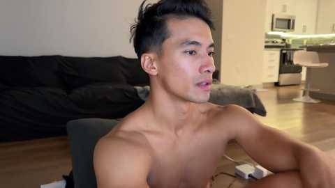 Media: Video of a shirtless, young, muscular Asian man with short black hair, sitting indoors, looking thoughtful. Background shows a dark sofa, kitchen with white counters, and a microwave.