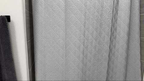 Media: Video of a bathroom shower curtain with a diamond-patterned design in shades of gray, hanging in front of a white wall. A dark gray towel is draped over a towel rack to the left.