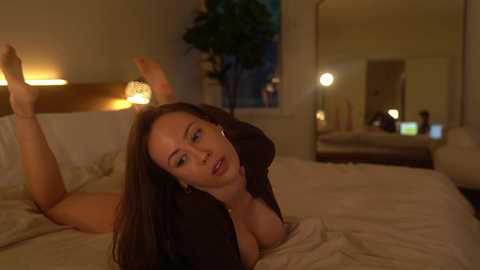 Media: Video of a fair-skinned woman with long brown hair, lying on a bed, wearing a black top, with a large mirror reflecting the room's warm lighting and a potted plant.