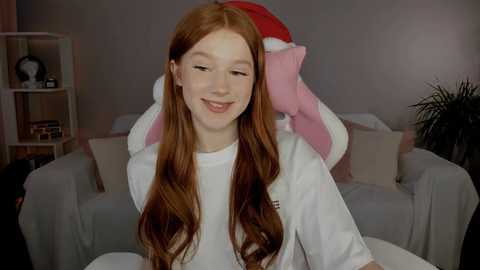 Video of a young red-haired girl with fair skin smiling in a cozy living room, wearing a white t-shirt. Behind her, a large plush toy dressed as a Christmas elf.