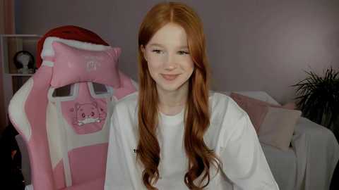 Video of a young woman with long, wavy red hair, smiling, wearing a white shirt, in a room with pink walls and a pink gaming chair with a cat pillow.