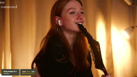 Media: Video of a fair-skinned, red-haired woman with long hair, wearing black lace gloves, singing into a microphone, with a blurred, warm-toned background.