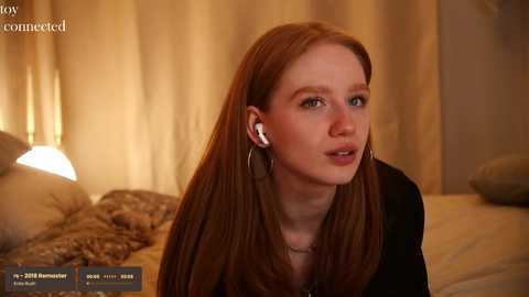 Media: Video of a fair-skinned, red-haired woman with straight hair, wearing a black top, sitting on a bed with beige sheets, talking on an earpiece. The background has beige curtains and a warm, dimly lit room.