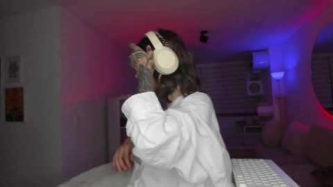 Media: Video of a person with headphones, wearing a white robe, hiding face, in a dimly lit room with purple and blue lighting, holding a laptop.