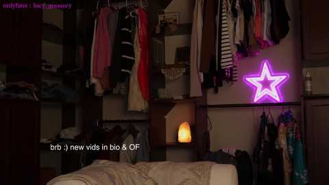 Media: Video of a messy, dimly-lit bedroom with clothes hanging on a rack, a glowing purple star-shaped neon light on the wall, and a small table lamp.