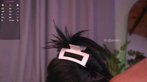 A close-up video of a person with dark hair styled into a black pompadour, adorned with a white plastic comb on the side, set against a blurred background with a green plant.