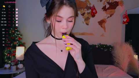 Media: A young woman with fair skin and brown hair, wearing a black choker, sucks on a yellow lollipop while standing in a dimly lit bedroom adorned with Christmas decorations.