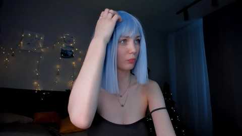 Media: Video of a pale-skinned woman with shoulder-length, straight, pastel blue hair, wearing a black strapless dress. She stands in a dimly lit room with string lights and posters on the wall.