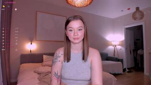 Media: Video of a young Caucasian woman with straight, shoulder-length brown hair, wearing a gray tank top, standing in a cozy, dimly-lit bedroom with beige walls, a bed, and a lamp.