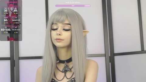 Video of a pale-skinned, slender woman with long, straight, platinum blonde hair, wearing a black choker and layered black necklaces. She has pointy elf ears and closed eyes. Background features a modern, minimalist room with a digital display showing stats.