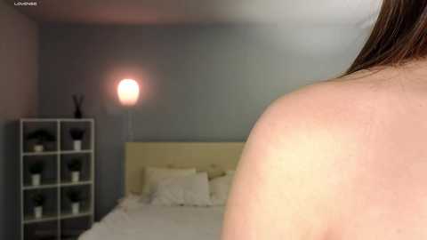 Media: A video shows a woman's bare back in a modern bedroom with a minimalist decor, featuring a white bed, a grey shelf with green potted plants, and a soft pink lamp.