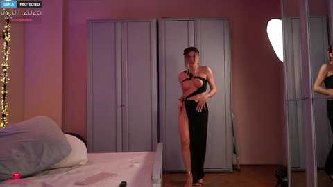 Media: A video shows a woman in a revealing black dress with a plunging neckline and thigh-high slit, standing in a dimly lit bedroom with a bed, wardrobe, and heart-shaped mirror.