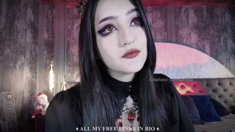 Media: Video of a pale-skinned woman with long black hair, wearing dark makeup and a gothic-inspired outfit, in a dimly lit, dark-themed bedroom with a plush bed and a teddy bear.