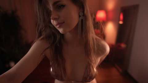 Media: Video of a young woman with long brown hair, wearing a low-cut, metallic-patterned bra, smiling slightly. Background features dimly lit, cozy room with red lamps, wooden furniture, and a blurred plant.