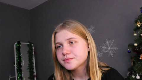 Media: Video of a young blonde girl with fair skin, wearing a black top, sitting indoors with a gray wall and Christmas decorations, including a tree and garlands.