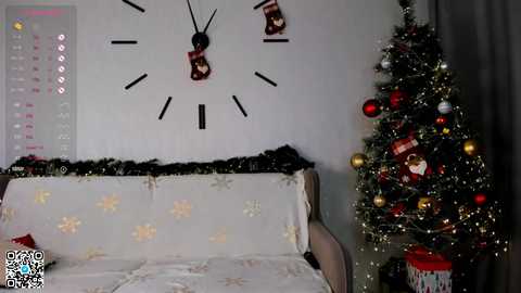 Media: Video of a cozy, festive bedroom with a white bed covered in snowflake-patterned sheets, a Christmas tree adorned with red ornaments, and a wall clock with a black face and red and white striped stockings.