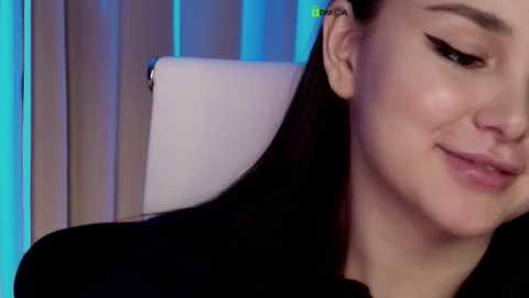 Close-up video of a smiling woman with long black hair, wearing a black top, against a blue-lit background.