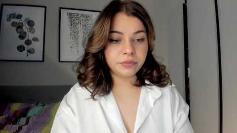 Media: Video of a young woman with wavy, shoulder-length brown hair, wearing a white robe, standing indoors with modern art on the wall in the background.