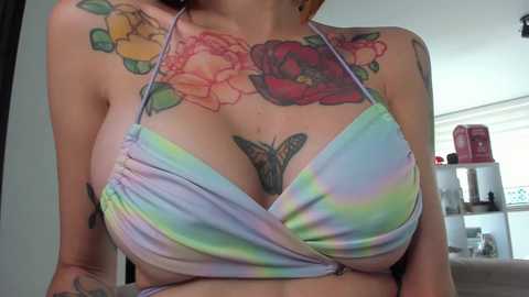 Media: Video of a light-skinned woman with colorful tattoos on her chest and shoulders, wearing a pastel tie-dye bikini top. Background includes a white wall and a bookshelf.