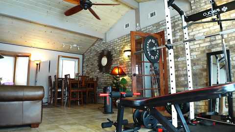 Media: Video of a rustic home gym with a brick wall, wooden ceiling, a large exercise bike, and a clock above a doorway.