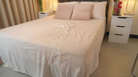 Media: Video of a neatly made bed with white sheets, two pink pillows, beige headboard, and a bedside table with a red flower vase.