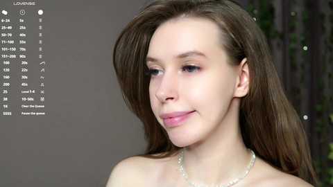 Media: Video of a young, fair-skinned woman with long brown hair, wearing a delicate necklace. She has a soft smile and a serene expression. Background is blurred, with a digital overlay of a calendar.