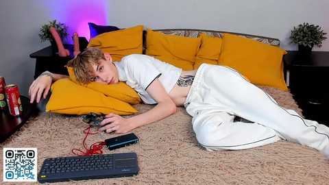 Media: Video of a young, slim, white boy with messy blond hair, lounging on a beige shag carpeted bed with mustard yellow pillows, wearing white athletic pants and a white shirt, holding a controller, surrounded by red and black gaming accessories.