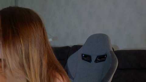 Media: Video of a woman with long, straight, light brown hair sitting on a dark gray, upholstered chair. A large, white, alien mask with black eyes and a wide, open mouth is placed on the backrest. The background is a plain, light-colored wall.