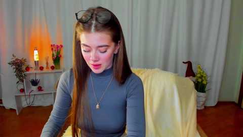 Media: Video of a young woman with long, straight brown hair, wearing a gray turtleneck, sitting on a chair in a softly lit room with white curtains, a lit candle, and floral decorations.