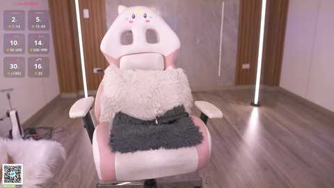 Media: Video of a plush pink and white cat-shaped gaming chair with a gray blanket, set against a minimalist, modern room with light wooden floors and glass doors.