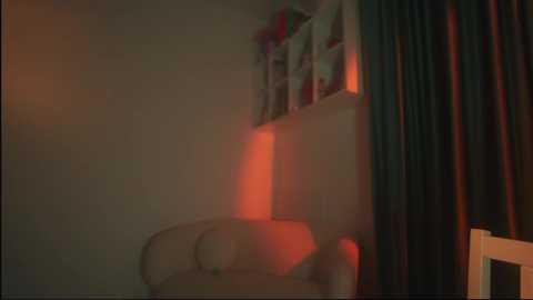 Media: A dimly lit video of a beige armchair, partially illuminated by a warm, red light, set against a dark, shadowy background with visible curtains and bookshelves. The overall ambiance is cozy yet slightly eerie.