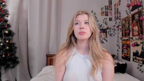 Media: Video of a young Caucasian woman with long blonde hair in a white tank top, standing in a festive, cluttered bedroom with a Christmas tree and wall posters.